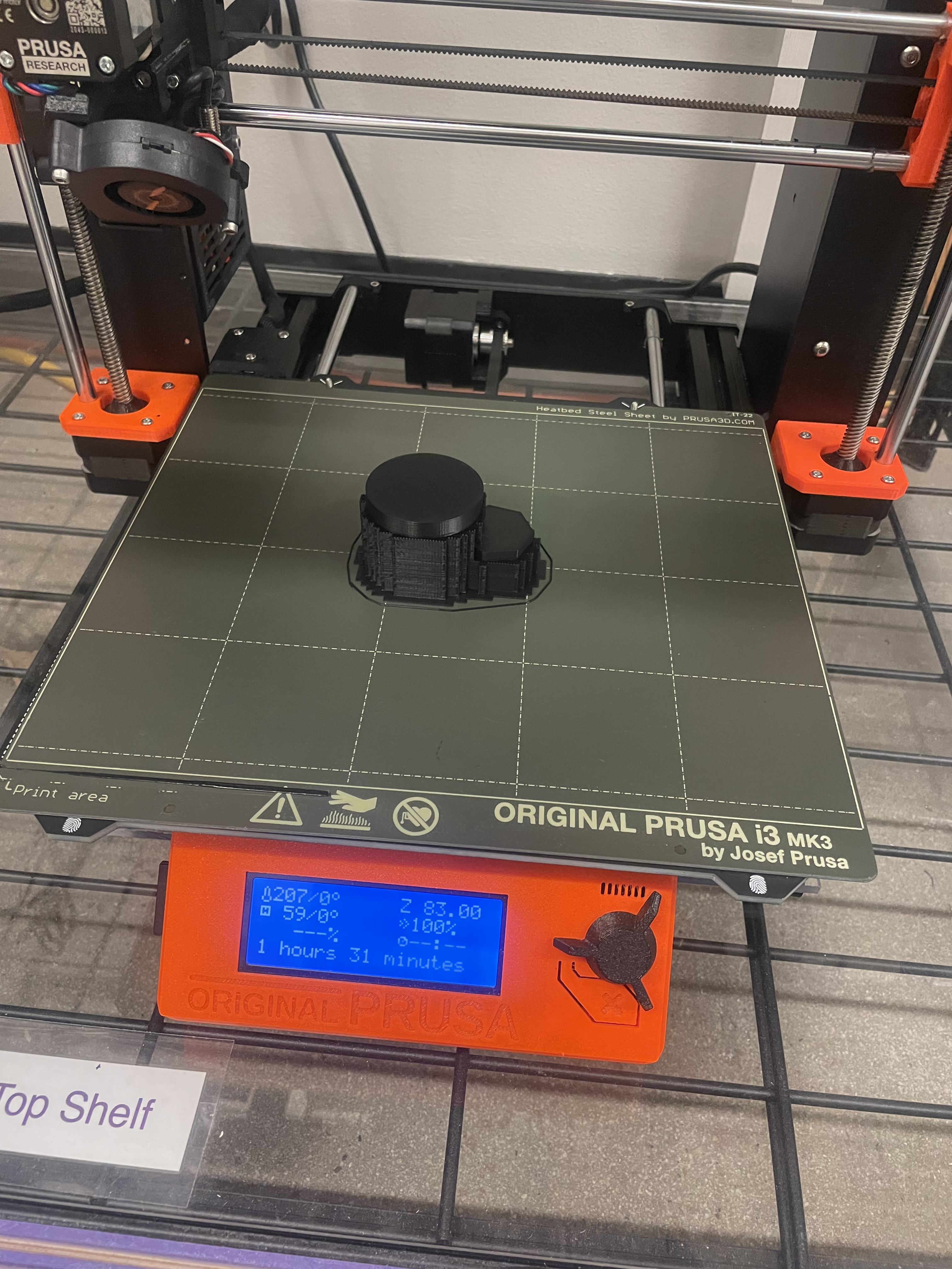 A Prusa MK3S+ printer with a completed project of black filament.  The project is surrounded by support material in a cube like shape.